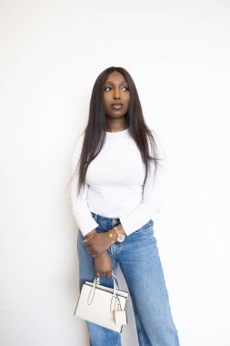 Beyaz bluzlu ve kot pantolonlu, şık bir tasarımcının el çantasını tutan, beyaz bir fonda poz veren kendine güvenen bir kadın..