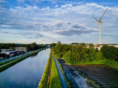 Halle, Belçika, 9 Temmuz 2024 'te bir kanal, rüzgar türbini, yeşillik ve mavi gökyüzünün altında yakalanan kırsal bir yol içeren sakin bir manzara..