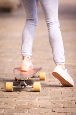 Tuğla bir yolda kaykay yapan biri, açık renk pantolon ve spor ayakkabılar giymiş, bacaklarına ve ayaklarına odaklanmış.