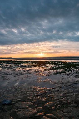 Sular çekildiğinde sakin bir sahil, yansıyan ışınlarla huzurlu bir gün batımı, dağınık bulutlar, sakin ve sakin bir atmosfer.