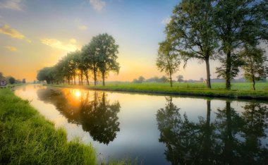 Serin yeşilliğin çerçevelediği sakin ve huzurlu bir nehri yansıtan büyüleyici bir gün batımı sahnesi.