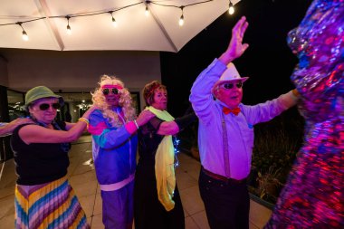 Particiler canlı kostümler içinde neşeli ışıklar altında dans ediyor, kutlama ve enerji gösterisi yapıyorlar.