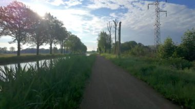 Sakin ve sakin bir patika, yemyeşil ve parlak mavi gökyüzü tarafından kucaklanan sakin bir kanal boyunca uzanır.