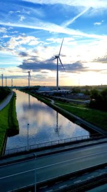 Güzel, resimli bir gün batımı sakin bir kanala yansıyor, sakin bir manzaranın ortasında rüzgâr türbinleri sergiliyor.