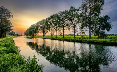 Gün batımında ağaçlarla çevrili güzel, resimli bir nehir manzarası canlı, gür yeşilliği yansıtıyor.
