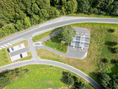 Güneş panelli büyük bir park alanını ve EV şarj istasyonunu güzel bir viraj yoluna bitişik olarak gösteren bir hava manzarası.