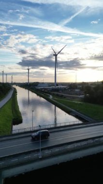 Güzel, sakin bir günbatımı manzarası bir nehir ve işlek bir otoyol kenarına yerleştirilmiş rüzgar türbinleri sergiliyor.