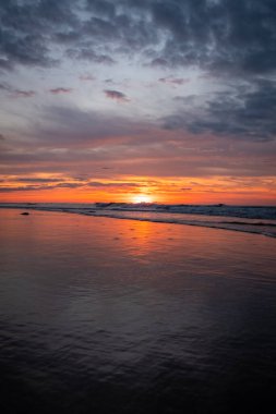 Gün batımında sakin plaj huzur yayar, gökyüzü sakin okyanus sularını yansıtır, barışçıl bir atmosfer yaratır.