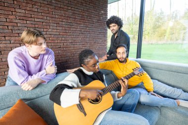 Bir grup arkadaş rahat, modern bir oturma odasında kanepede dinlenirken, bir tanesi akustik gitar çalıyor.