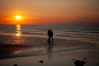 Huzurlu, huzurlu bir plaj sahnesinde çarpıcı bir günbatımının keyfini çıkarırken bir çift birbirini neşeyle kucaklıyor.