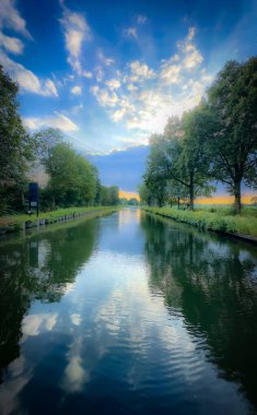 Durgun bir nehir, her iki tarafında yemyeşil yemyeşil bulutlarla çevrili, parlak gökyüzünü yansıtır.