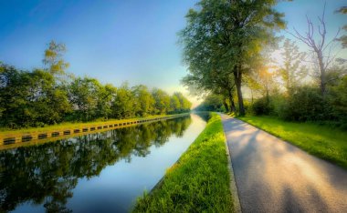 Berrak mavi gökyüzünün altında yemyeşil ağaçlarla kaplı güzel bir kanal yolunun huzurlu ve huzurlu manzarası.