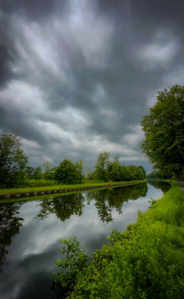 A tranquil river scene reflecting dark, ominous clouds that loom above amidst the lush, vibrant greenery surrounding it