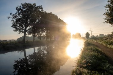 Sis ve yemyeşil ağaçlarla kaplanmış, gün ışığını güzel yansıtan sakin bir sabah sahnesi.