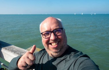 Neşeli bir adam, bu güzel sahil kenarında, güneşli ve aydınlık bir günün tadını çıkarırken, coşkulu bir şekilde başparmağını kaldırır.