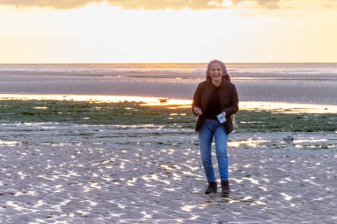 Genç bir kadın gün batımında sahilde huzur içinde yürüyor. Etrafı sakin sular ve güzel bir gökyüzü ile çevrili.