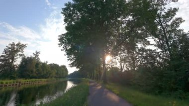 Huzurlu ve huzurlu bir yol, zarif bir şekilde uzun ağaçlarla kaplı güneşli bir günde dingin bir kanalın yanında uzanır.