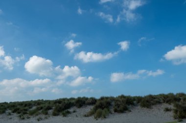 Kumulların, parlak mavi gökyüzünün ve üzerinde yavaşça süzülen kabarık bulutların olduğu huzurlu bir sahil manzarası.