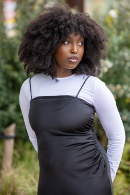 Doğal kıvırcık saçlı, siyah elbiseli, beyaz bluzlu, açık havada poz veren şık bir kadın..