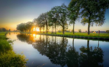 Güzel bir günbatımı, gür ağaçlar ve canlı renklerle çevrili sakin bir nehrin üzerine sıcak bir ışık saçar.