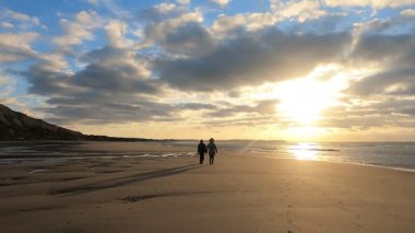 Huzurlu ve huzurlu bir sahil sahnesi. Baş döndürücü bir gün batımında bir çift el ele yürüyor.