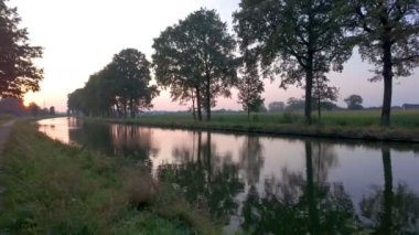 Gün batımının canlı renklerini yansıtan gür ağaçlar ve dingin bir kanal içeren nefes kesici ve resimsel bir sahne.