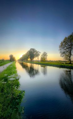 Huzurlu ve huzurlu bir nehir manzarası, güzel yansımaları olan, bereketli bir günbatımı gökyüzünün altında, yemyeşil ağaçlarla çevrili