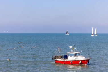 Oostend, Belçika, 6 Ekim 2024, parlak ve canlı kırmızı bir balıkçı teknesi sakin suların ve ufuktaki uzak yelkenlilerin arasında duruyor.