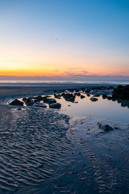Güzel, huzurlu bir gün batımı kayalık bir sahili aydınlatıyor. Dalgalar her tarafı canlı renklerle yansıtıyor.