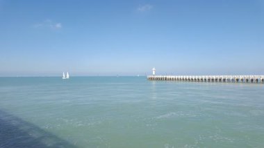 Nieuwpoort, Batı-Flanders, Belçika, 6 Ekim 2024 çarpıcı güzellikteki sahil manzarasında zarif yelkenliler, tarihi bir deniz feneri ve açık mavi gökyüzü bulunur.