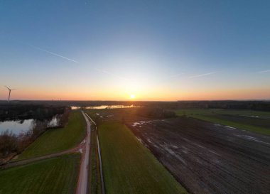 Göz kamaştırıcı bir günbatımı, yemyeşil tarlaların üzerine ılık bir parıltı ve sakin bir su yolu sergiliyor.