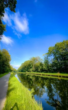 Huzurlu ve sakin bir kanal sahnesinde berrak yansımalar, yemyeşil bankalar ve yukarıda parlak mavi gökyüzü sergilenir.