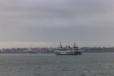 Den Helder, Hollanda, 28 Ekim 2024, yoğun bir limana yaklaşan bir feribot parçalı bulutlu bir gökyüzü altında hareketli deniz aktivitelerini vurguluyor.