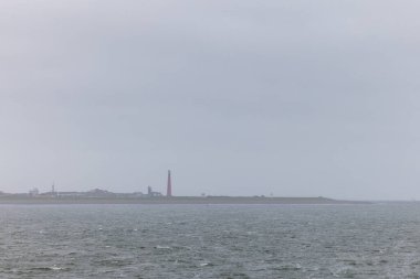 Den Helder, Hollanda, 28 Ekim 2024, sakin bir kıyı manzarası karamsar, bulutlu bir gökyüzünün altında güzel bir şekilde çerçevelenmiş uzak bir deniz fenerini gösteriyor.