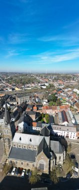 Lembeek, Halle, Vlaams Brabant, Belçika, 4 Mart 2025, çarpıcı bir hava manzarası, berrak mavi gökyüzünün altındaki canlı bir şehir manzarası tarafından çerçevelenmiş antik bir kiliseyi gösteriyor.