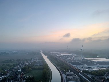 Lot, Beersel, Vlaams-Brabant, 22 Nisan 2025, sisli bir manzaranın ortasında şafak vakti rüzgar türbinlerinin eşlik ettiği dolambaçlı bir nehrin sakin ve sakin bir görüntüsü.