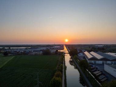 Rijkevorsel, Antwerp, Belçika, 27 Nisan 2025, çarpıcı bir günbatımı yeşil alanlar ve sanayi binaları yakınlarındaki sakin bir kanala yansıyarak büyüleyici bir manzara yaratıyor.