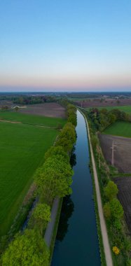 Sint Jozef, Rijkevorsel, Antwerp, Belçika, 27 Nisan 2025, gün batımında yeşil tarlalarda gezinen huzurlu bir nehri görüntüleyen bir görüntü.