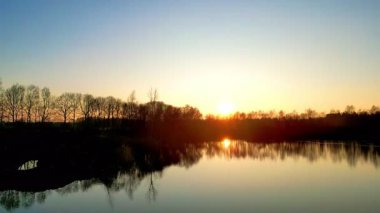 Sakin bir gündoğumu, ağaçlar ve doğa tarafından mükemmel çerçevelenmiş durgun suları güzel bir şekilde yansıtır.