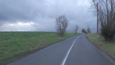 Huzurlu ve sakin bir köy yolu, gür tarlalar ve uzun ağaçlar arasında kıvrılıyor, hepsi kasvetli bir gökyüzüne karşı kurulmuş.