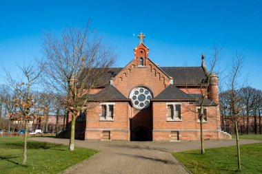 Chapel, Kolonie, Merksplas, Antwerp, Belçika, 26 Nisan 2025 çarpıcı kırmızı tuğla bir kilise güzel dairesel bir pencereye sahiptir, bereketli ağaçlar ve parlak mavi gökyüzü tarafından zarif çerçevelenmiştir.