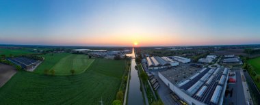 Rijkevorsel, Antwerp, Belçika, 27 Nisan 2025 'te güzel bir gündoğumu sanayi bölgelerine altın ışık saçtı ve dingin bir su yolu kenarında yemyeşil alanlar oluştu.