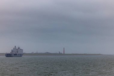 Den Helder, Hollanda, Ekim 28, 2024, yoğun sisli sularda ilerleyen büyük bir konteyner gemisi bulutlu gökyüzünün altındaki sanayi kıyı şeridine yakın.