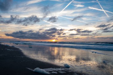 Çarpıcı bir sahil gün batımı dramatik bulutların ve çizgili gökyüzünün altındaki yumuşak dalgaları güzel bir şekilde yansıtıyor.