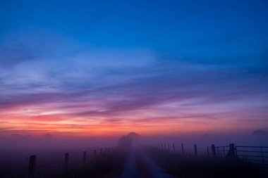 Hafif sisli bir sabah yolu kırsalda canlı ve renkli bir gündoğumuyla güzelce aydınlatılır.