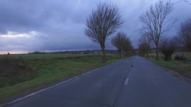 Bereketli ağaçlarla kaplı dolambaçlı, sakin bir kır yolu manzarası dramatik, karamsar, bulutlu bir gökyüzüne kurulmuş.
