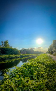 Bereketli yeşilliğin ve yukarıda parlayan parlak güneşin yer aldığı sakin ve sakin bir nehir manzarası güzellikleri gözler önüne seriyor.