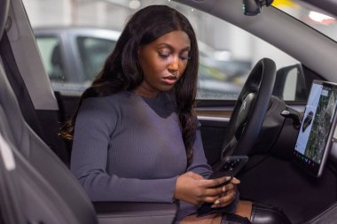 Afro-Amerikalı bir kadın şık bir iç mimarisi olan modern bir arabanın sürücü koltuğunda otururken akıllı telefonuna odaklanır. Gösterge panelinde geniş bir dokunmatik ekran görüntüsü var.