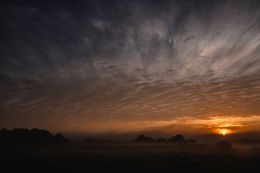 Bu ilgi çekici görüntü alacakaranlığın yoğun ve düşünceli güzelliğini gözler önüne seriyor. Karanlık, engin gökyüzü dramatik bulut oluşumlarıyla çizilirken, batan güneş ateşli bir odak noktası oluşturuyor.