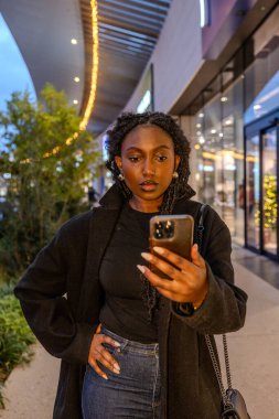 Şık küpelerle süslenmiş ve saçları dar kıvrımlı genç bir siyahi kadının şehir alacakaranlığı ortasında akıllı telefonuyla bir anlık bağlantı kurmasıyla ilgi çekici bir sahne ortaya çıkıyor. Şey...